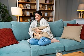 Das Bild zeigt eine Frau, die mit Bauchschmerzen auf der Couch sitzt.